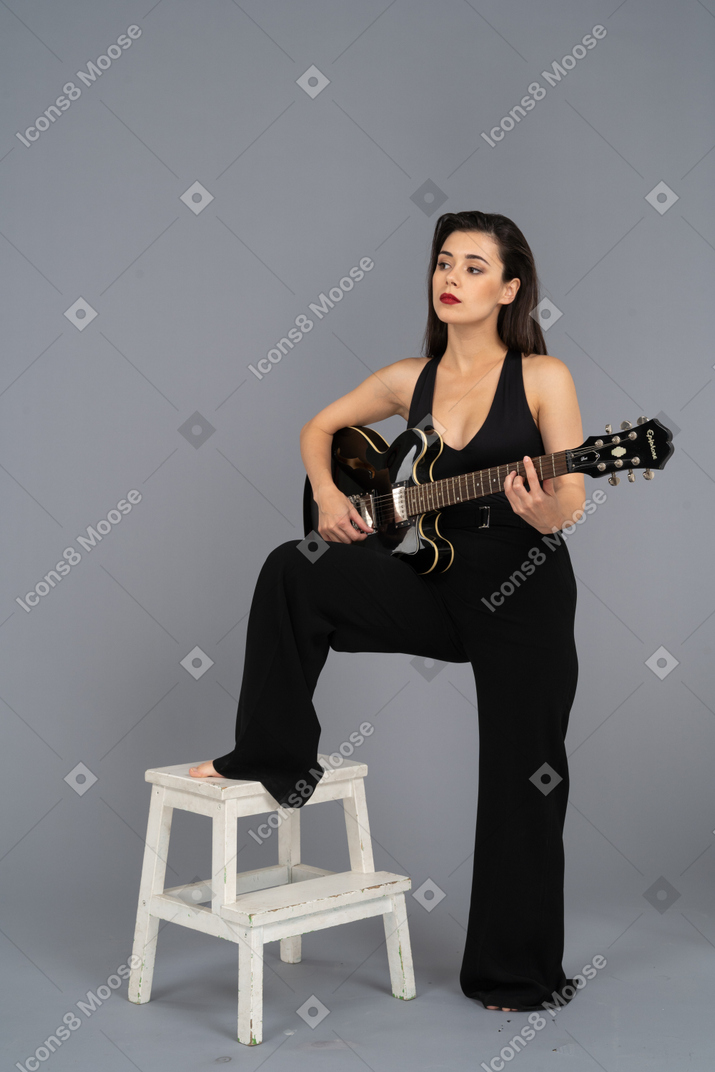 기타를 들고 의자에 다리를 씌우고 검은 양복을 입은 젊은 아가씨의 3/4보기