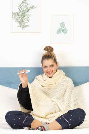 Vista frontal de una joven envuelta en una manta blanca sentada en la cama con termómetro