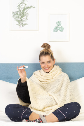 Front view of a young lady wrapped in white blanket sitting in bed with thermometer