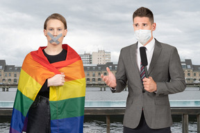 Reporter in face mask talking to a young woman with taped mouth holding a pride flag 