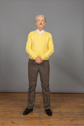 Vue de face d'un vieil homme en pull jaune tenant les mains avec ses yeux fermés