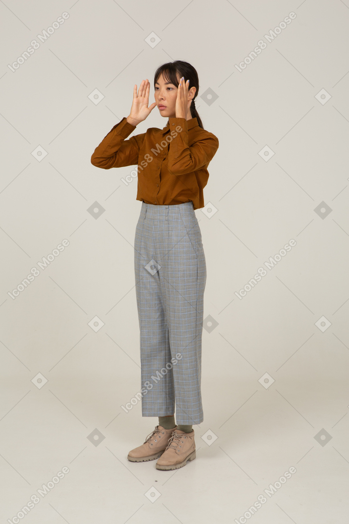 Vista di tre quarti di una giovane donna asiatica in calzoni e camicetta che alza le mani