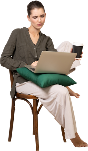 당황한 젊은 여성이 의자에 앉아 노트북과 커피 컵을 들고 있는 모습