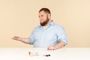 Hombre grande sentado a la mesa y mostrando esmaltes de uñas