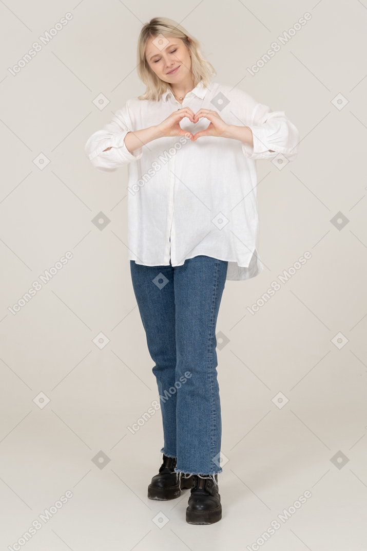 Front view of a blonde female in casual clothes showing a heart gesture with her eyes closed