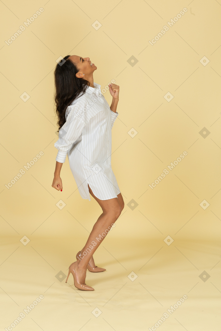 Side view of a dancing dark-skinned young female in her white dress raising hands