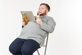 Bid bearded man holding laptop on belly, watching movie and having chips and beer