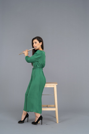 Вид сбоку на девушку в зеленом платье, сидящую на стуле и играющую на кларнете