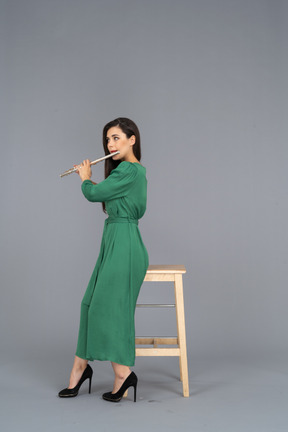 Side view of a young lady in green dress sitting on a chair while playing the clarinet