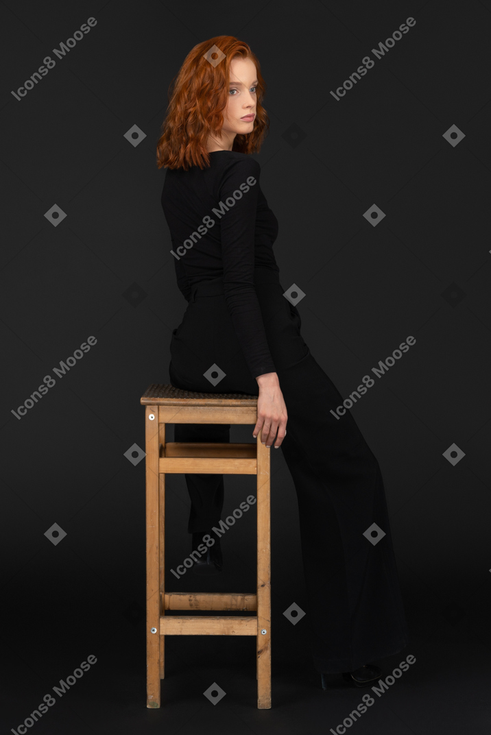 A back side view of the cute young woman sitting on the wooden chair and looking to the right