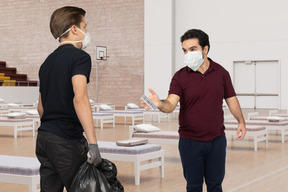 Man in face mask holding a trash bag standing next to a man in face mask with measuring device at the hospital