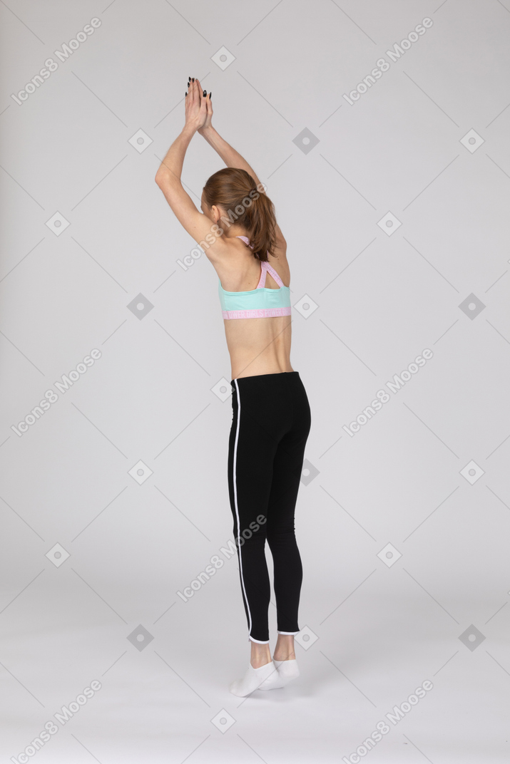 Vista posterior de tres cuartos de una jovencita en ropa deportiva de pie de puntillas y levantando las manos