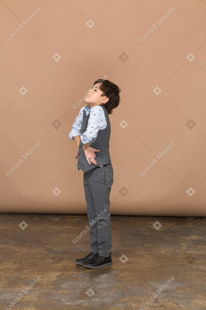 Вид сбоку на симпатичного мальчика в костюме, стоящего с руками на бедрах