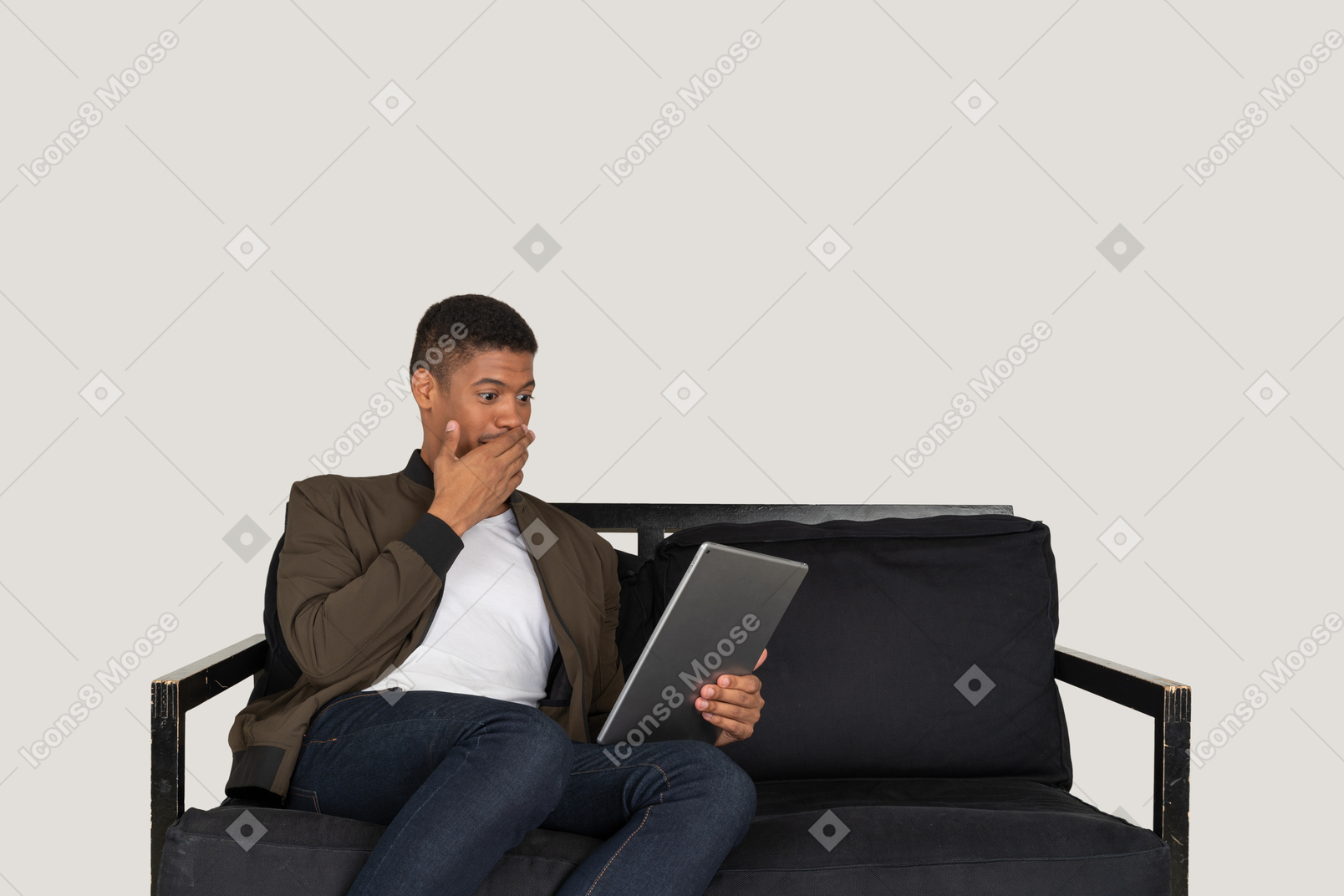 Front view of a shocked young man sitting on a sofa while watching the tablet