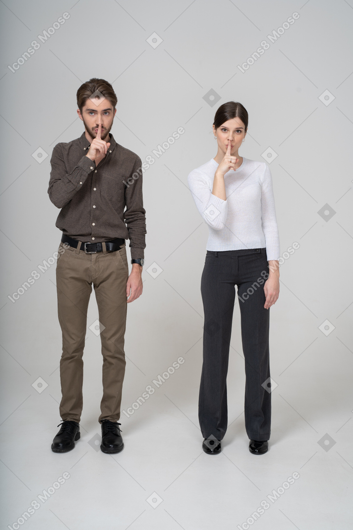 Vista frontal de um jovem casal com roupa de escritório mostrando gesto de silêncio