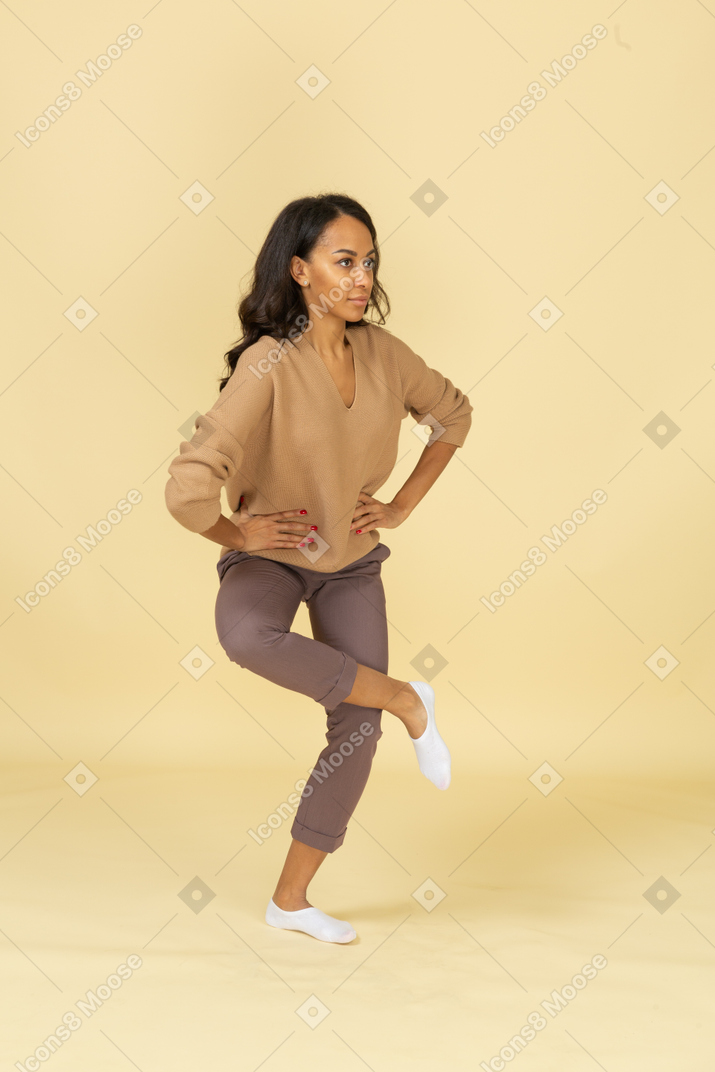 Вид в три четверти темнокожей молодой женщины, поднимающей ногу и кладущей руки на бедра