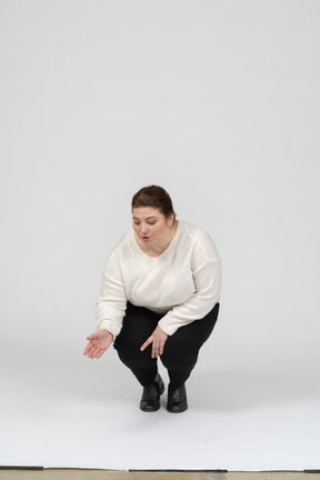 Женщина большого размера в белом свитере сидит на корточках, вид спереди