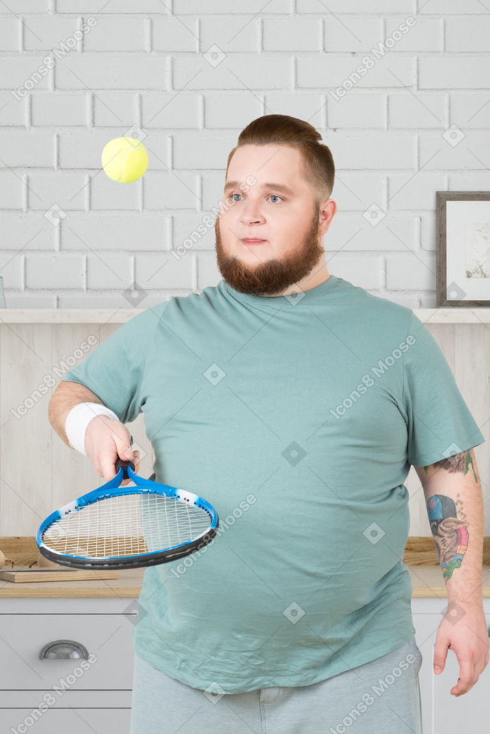 A fat man holding a tennis racket and playing with a tennis ball