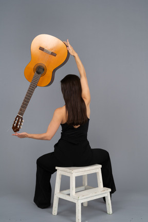 Vista posterior de una joven en traje negro sosteniendo la guitarra sobre la cabeza y sentada en un taburete