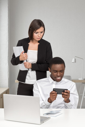 A female boss looking over shoulder of man playing with smartphone at the office