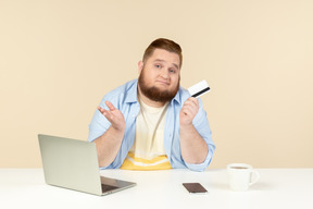悲伤看起来年轻超重的男人坐在办公桌前，拿着银行卡