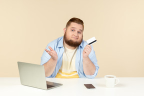 Triste olhando jovem homem com excesso de peso, sentado na mesa e segurando o cartão do banco