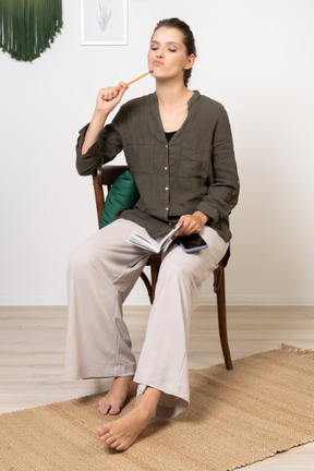 Vorderansicht einer nachdenklichen jungen frau in hauskleidung, die mit bleistift und notizbuch auf einem stuhl sitzt