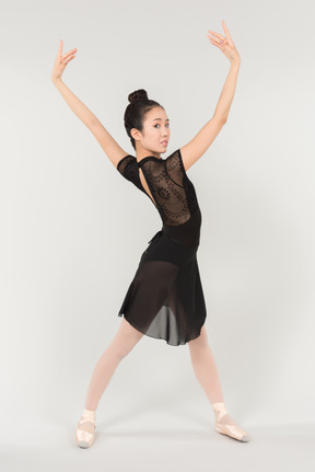 Молодая азиатская балерина стоит с поднятыми руками и наполовину сбоку от камеры