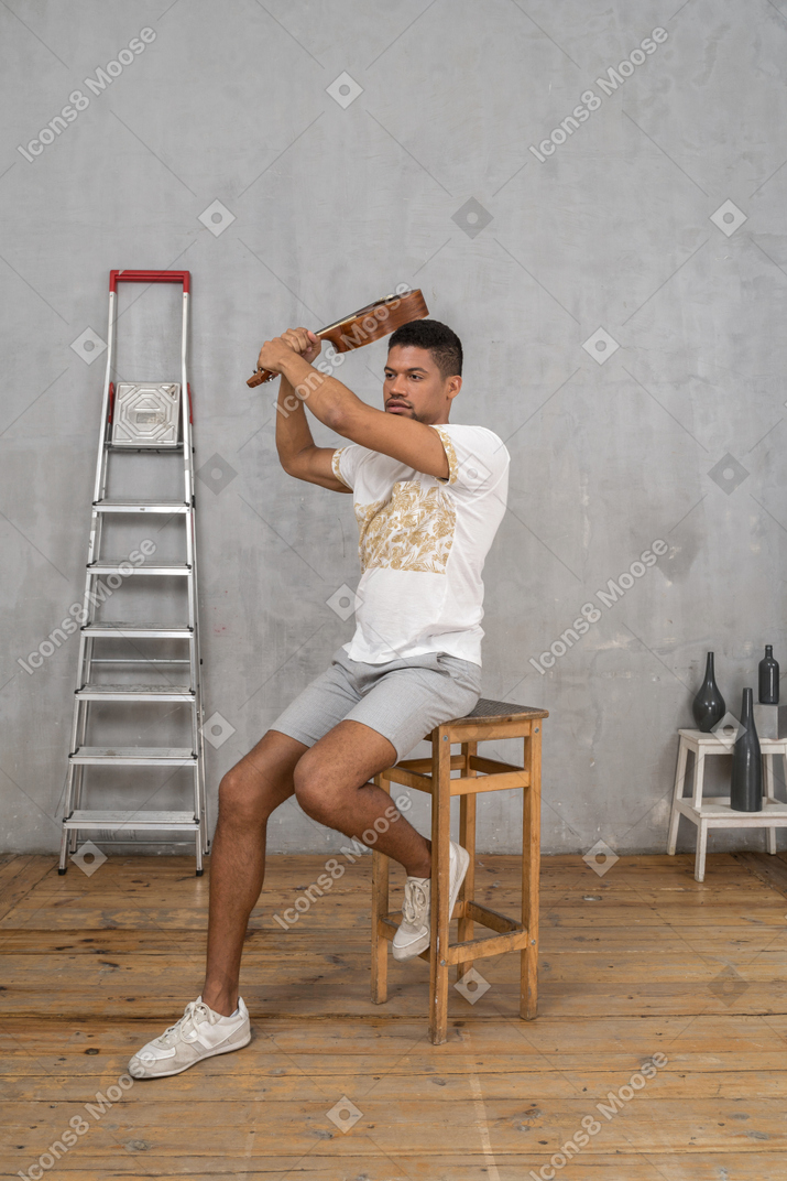 Three-quarter view of a man on a stool swinging a ukulele like a bat