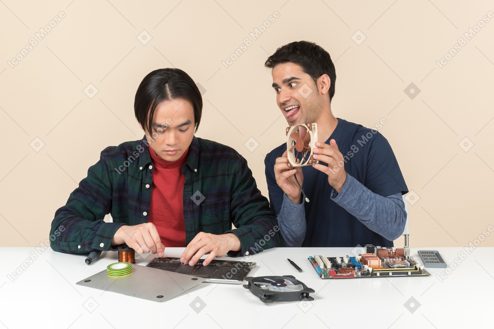 Two young geeks sitting at the table with details on it and one of them is fooling around