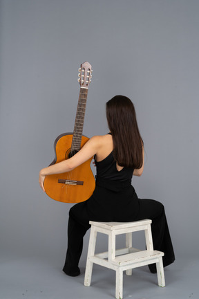 Vista posterior de una joven en traje negro sosteniendo la guitarra y sentada en un taburete