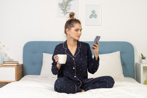 Devant une jeune femme en pyjama assise dans son lit avec une tasse de café tout en vérifiant instagram