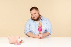 Lächelnd schüchterner junger großer mann, der am tisch sitzt und barbie-puppe hält