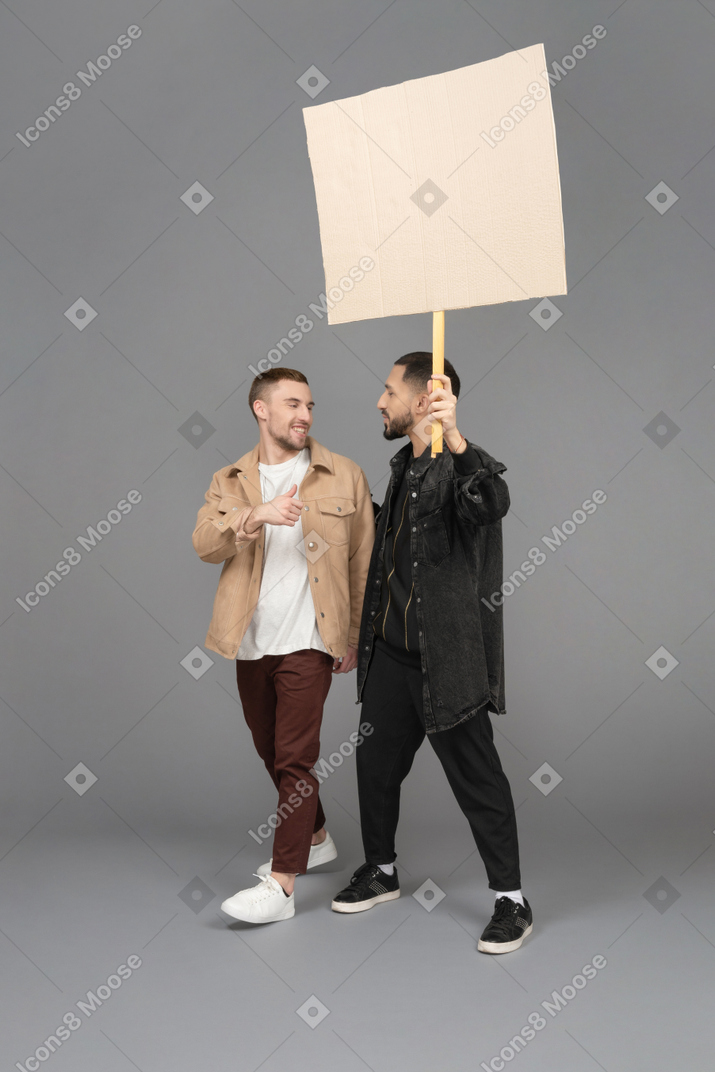 Vista de tres cuartos de dos hombres jóvenes que llevan una valla publicitaria y charlan