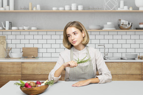 A woman sitting at a table with a bowl of vegetables and holding herbs