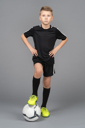 Vista frontal de un niño chico en uniforme de fútbol poniendo las manos en las caderas y el pie en la bola