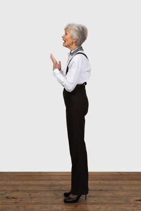 Vista laterale di una donna anziana contenta vestita in abiti da ufficio mettendo le mani insieme aprendo la bocca e gesticolando