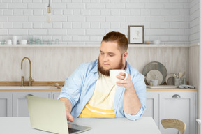 A bearded man sitting at a table with a laptop and a cup of coffee