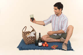 Young caucasian man holding glass of wine while sitting on the blanket