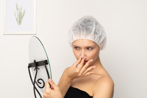 Mujer infeliz con gorra quirúrgica mirándose en el espejo y tocándose la nariz