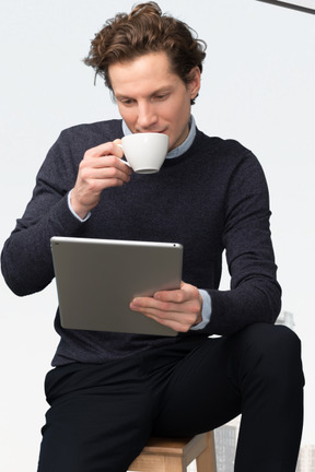 A man sitting on a stool drinking a cup of coffee while using a tablet