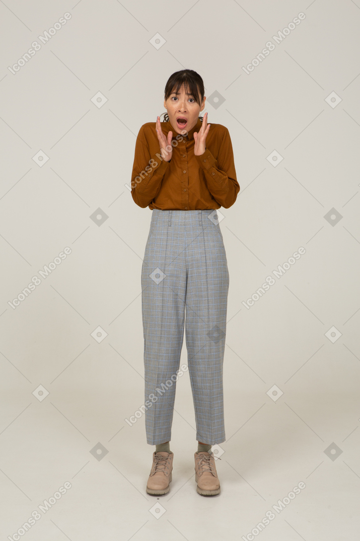 Front view of a shocked young asian female in breeches and blouse raising hands