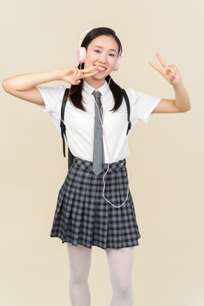 Asian school girl listening to music in headphones and showing v sign