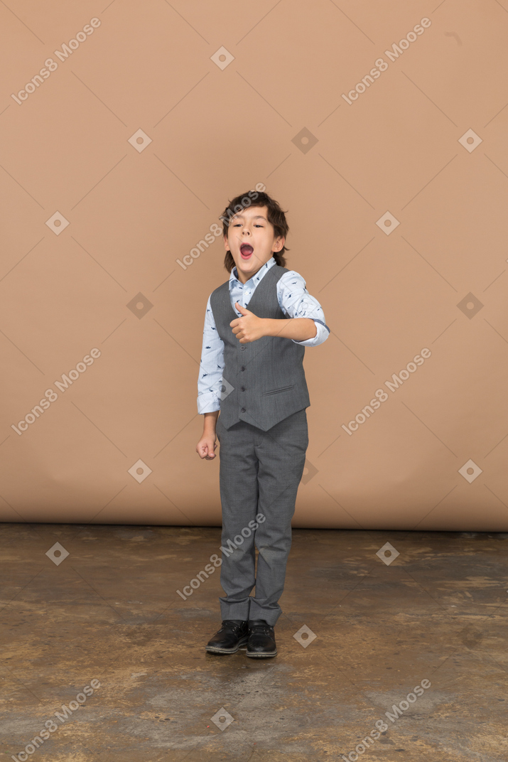Vue de face d'un garçon mignon en costume gris montrant le pouce vers le haut