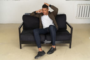 Front view of a perplexed young man sitting on a sofa and talking on his phone