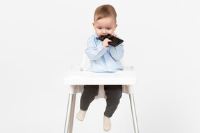 Baby boy sitting in highchair and bitting a mobile phone