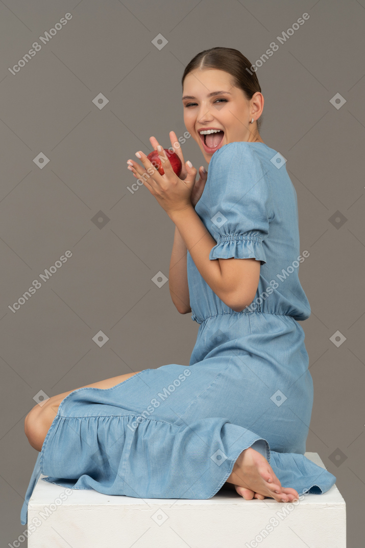Side view of cheerful young woman with apple sitting on cube and looking at camera