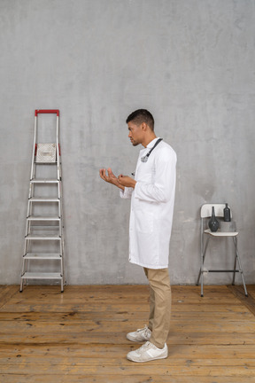 Vista lateral de un joven médico de pie en una habitación con escalera y silla explicando algo