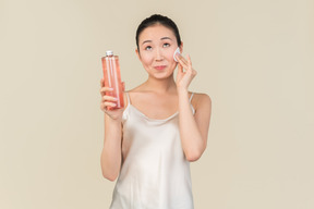 Dreamy young asian girl holding cosmetic bottle and cotton pad