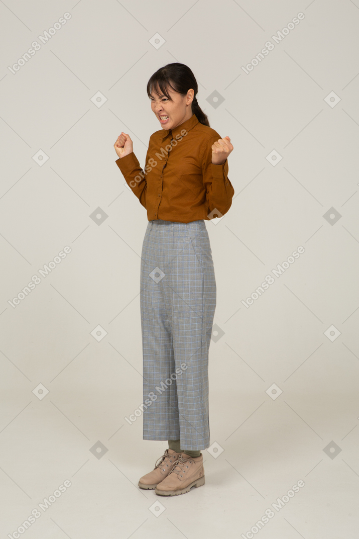 Vista de tres cuartos de una joven mujer asiática malvada en calzones y blusa apretando los dientes y los puños